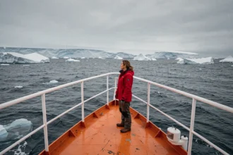 Voyageuse en parka rouge à la proue d'un navire d'expédition en Antarctique face aux icebergs